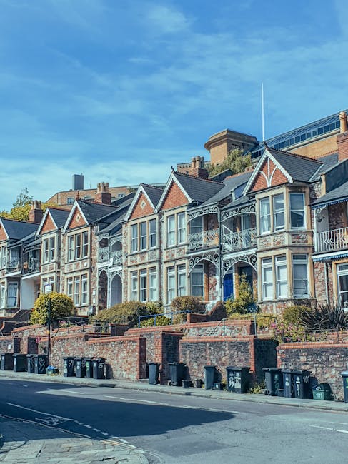 A row of Victorian-style terraced houses with ornate facades, bay windows, and decorative metalwork on balconies, situated on a residential street during daytime with clear skies. In the foreground, a pavement and curbline are visible, with several black and green wheelie bins lined up along the sidewalk. The houses are elevated above the street level, accessed by short brick stairs with metal railings. Behind the houses, there is a larger building with a glass roof, indicating a mix of architectural styles. The scene suggests a quiet urban area suitable for professional house removals and home relocation services, with Movers Elephant and Castle providing detailed packing and furniture transport during the loading process.