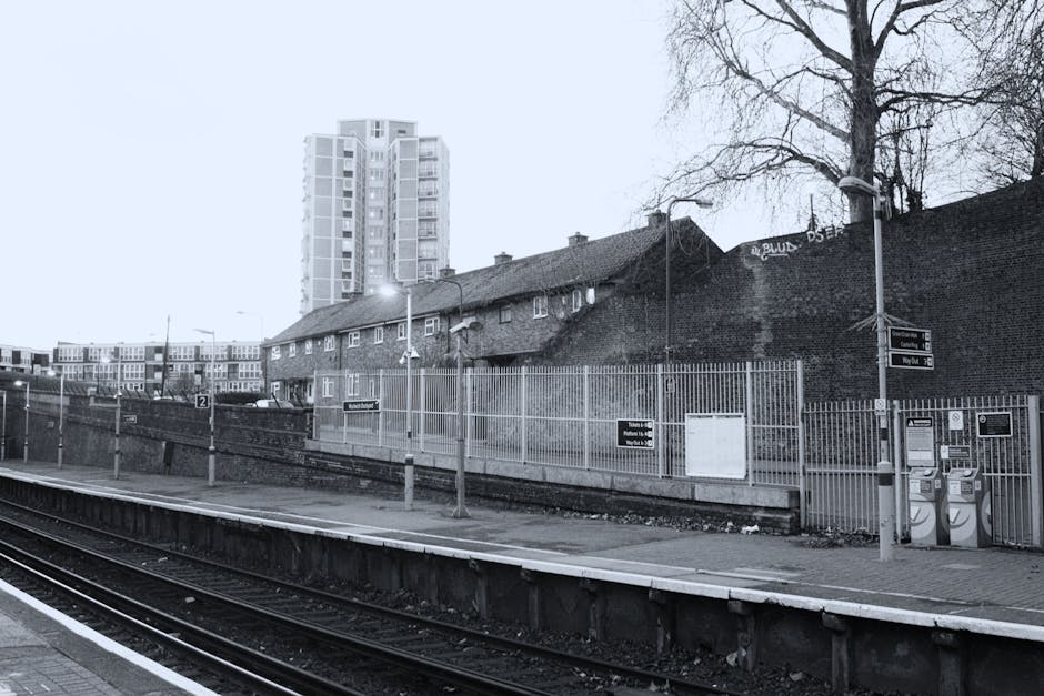 A black and white photograph of Elephant and Castle railway station platform showing metal train tracks in the foreground, with a tiled platform edge and a small set of electric vehicle charge points on the right side. Behind the platform, there is a tall brick wall with some graffiti and a leafless tree extending above it, indicating wintertime. Further back, a row of wooden residential houses with pitched roofs and small windows are visible, set against a backdrop of modern high-rise apartment buildings with multiple windows and balconies, under an overcast sky. Street lamps are mounted on poles along the platform, illuminating the area with soft light. The environment appears quiet and devoid of passengers, with a focus on the station infrastructure and surrounding urban landscape. This image is associated with house removals, reflecting the logistics involved in furniture transport and home relocation services by Movers Elephant and Castle.