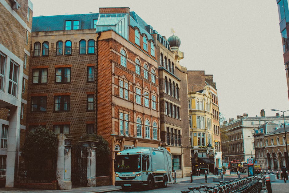 Photograph of a city street scene in Elephant and Castle showing a white moving van positioned on the pavement beside historic multi-storey buildings constructed from red and brown brick, featuring large arched windows and decorative facades. The van appears to be part of a home relocation or furniture transport operation by Movers Elephant and Castle, with the vehicle’s back door open for loading or unloading furniture and packing materials. The sidewalk is lined with metal bike racks, and there are various buildings with architectural details such as cornices and parapets in the background. The street is illuminated by street lamps, and a few pedestrians and other vehicles are visible in the distance, indicating active urban movement. The scene captures the process of packing, loading, and transporting household or office furniture as part of a professional removals service in an active city environment.