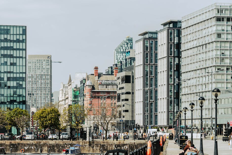A cityscape featuring modern office buildings with glass facades and contemporary architectural designs, situated along a busy street in Elephant and Castle. In the foreground, there is a paved pedestrian area with black metal lampposts lining the sidewalk, where a woman sits on a bench. Several trees with green foliage are planted along the pavement, separating the walkway from the road. Parked cars and vans are visible along the curb, including a large white van likely used for home relocation or furniture transport, connected to the services of Movers Elephant and Castle. The scene captures the loading process of packing and moving activities, with some motion implied by the positioning of the van near a building entrance, possibly indicating loading or unloading of boxes, furniture, or packing materials such as cardboard boxes, plastic wrap, and blankets. The overall environment reflects an urban setting in the midst of a professional moving operation, with a clear daylight sky overhead providing natural lighting.
