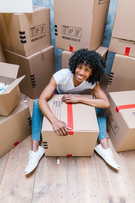 A man with dark curly hair and a beard is inside a bright, empty room with white walls and wooden flooring, engaged in a home relocation process. He is dressed in casual dark clothing, including a t-s