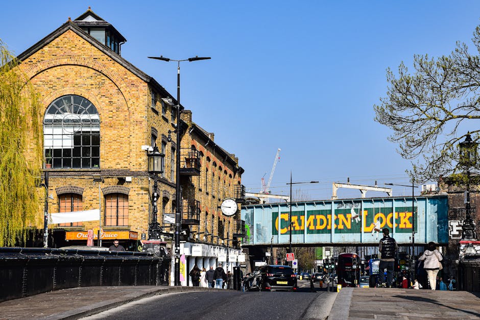 A street scene in Elephant and Castle featuring a brick building with large arched windows and a clock mounted on a lamppost. In the foreground, a loading area is visible with a moving company van belonging to Movers Elephant and Castle. The van is positioned near the curb, and several cardboard boxes, some wrapped in plastic or fabric, are being loaded or unloaded, indicating moving or packing activities. Nearby, people are walking along the pavement, and a trolley with additional boxes is seen near the entrance of the premises. Overhead, streetlights and a railway bridge with a painted sign reading 'Caidin Lock' span across the street, with some urban construction cranes visible in the background under a clear blue sky. The environment is well-lit, highlighting the details of the furniture, packaging materials, and the busy moving process associated with home relocation services.