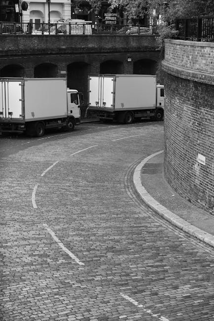 A large commercial delivery truck with a green cargo area is parked on an urban street in front of modern and historic buildings in the city of London. The truck is positioned close to the curb, with its rear doors closed, and appears to be in the process of loading or unloading. The street features a paved surface with some pedestrians visible walking nearby, and the surrounding architecture includes a tall glass skyscraper reflecting the sky and other buildings, alongside older stone-faced structures with multiple windows. Bright daylight illuminates the scene, casting shadows of the truck and nearby buildings, and the overall environment suggests a typical city centre setting suitable for home or office relocations conducted by professional moving services, such as Movers Elephant and Castle, specializing in house removals and furniture transport in the SE1 area.